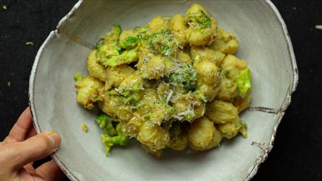 A bowl of creamy broccoli gnocchi mixed with broccoli and topped with grated cheese. The dish appears to be seasoned with herbs and has a creamy texture. A hand is holding the bowl from the side on a dark surface.